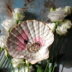 Könnte beinhalten: Eine weiße Keramik-Schmuckschale mit goldfarbenem Rand und floralem Design. Die Schale zeigt rosa Blumen mit handschriftlichem Text in schwarzer Tinte. Die Schale ist mit goldenen Ringen gefüllt.