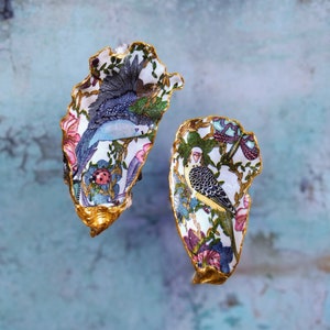 Könnte beinhalten: Zwei Austernmuscheln, die mit einem floralen Muster und Vögeln bemalt sind. Die Muscheln sind vergoldet und haben einen weißen Hintergrund mit bunten Blumen, Blättern und Vögeln.
