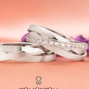 Könnte beinhalten: Zwei silberne Eheringe mit einem Diamantenband oben. Die Ringe sind ineinander verschlungen und haben eine polierte Oberfläche. Das Bild ist auf einem rosa Hintergrund mit dem Text "GÜMÜŞÇÜM" unten.
