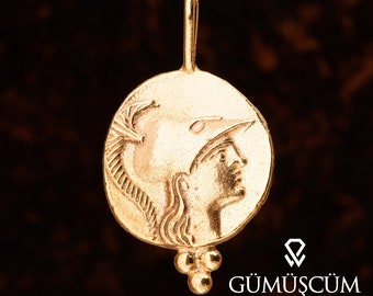 May include: A gold pendant necklace featuring a round coin with a detailed profile of a woman wearing a helmet. The coin is attached to a chain with three small gold beads at the bottom.