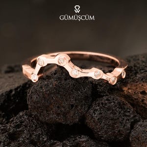 Könnte beinhalten: Roségoldener Ring mit einem einzigartigen Wellendesign, besetzt mit einer Reihe kleiner, klarer Edelsteine. Der Ring wird auf einer dunklen, strukturierten Oberfläche präsentiert. Der Markenname "GÜMÜŞCÜM" ist sichtbar.