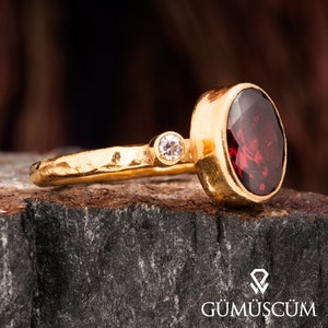 Könnte beinhalten: Ein goldener Ring mit einem großen, ovalen, roten Edelstein und einem kleineren, runden, klaren Edelstein. Die Ringband ist strukturiert.