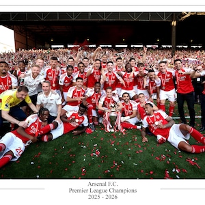 May include: A group of people in red and white soccer uniforms celebrate a victory, holding a trophy. The image includes the text "Arsenal F.C. Premier League Champions 2025 - 2026". Confetti is scattered on the green field.