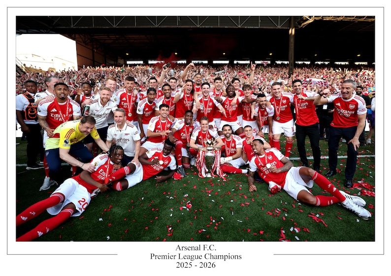 May include: A group of people in red and white soccer uniforms celebrate a victory, holding a trophy. The image includes the text "Arsenal F.C. Premier League Champions 2025 - 2026". Confetti is scattered on the green field.