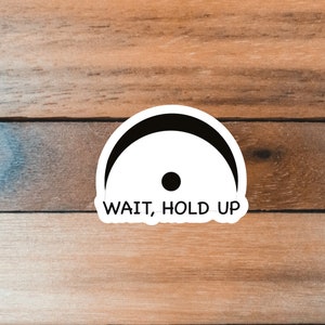Pu&ograve; includere: Un adesivo bianco con un semicerchio nero e un punto nero, con la scritta "WAIT, HOLD UP". L'adesivo &egrave; su una superficie di legno.