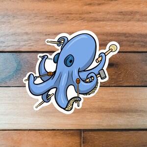 May include: A blue octopus with eight tentacles holding various percussion instruments, including mallets, drumsticks, and a small drum. The octopus is on a wooden background.