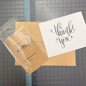Puede incluir: Un sobre marr&oacute;n con una tarjeta blanca en su interior. La tarjeta tiene una caligraf&iacute;a negra que dice "gracias". Una bolsa de pl&aacute;stico transparente con una tarjeta marr&oacute;n en su interior. La tarjeta tiene un logotipo negro que dice "MLD Melody Line Designs" y el texto "Instagram @melodylinedesigns Enjoy!"