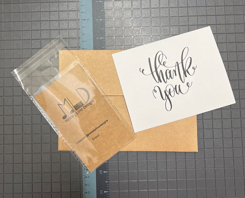 Puede incluir: Un sobre marr&oacute;n con una tarjeta blanca en su interior. La tarjeta tiene una caligraf&iacute;a negra que dice "gracias". Una bolsa de pl&aacute;stico transparente con una tarjeta marr&oacute;n en su interior. La tarjeta tiene un logotipo negro que dice "MLD Melody Line Designs" y el texto "Instagram @melodylinedesigns Enjoy!"