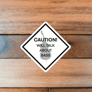 May include: A white diamond-shaped sticker with black borders and a gray silhouette of a bass guitar. The sticker reads "CAUTION! WILL TALK ABOUT BASS".