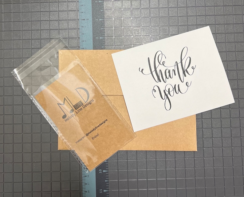 Pu&ograve; includere: Un sacchetto di plastica trasparente contenente una carta marrone con un logo e testo, accanto a una busta marrone e una carta bianca con le parole "thank you" in scrittura nera. Lo sfondo &egrave; una superficie grigia con un motivo a griglia.