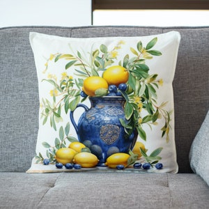 Puede incluir: Una almohada blanca con una jarra de cerámica azul y blanca llena de limones y hojas verdes. La jarra tiene un diseño floral y está rodeada de limones y arándanos.