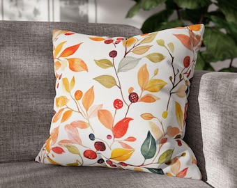May include: Square cushion featuring a watercolour design of autumn leaves and berries in shades of orange, red, yellow, and green on a white background. The cushion is on a grey sofa.