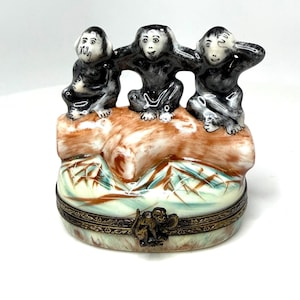 May include: A decorative trinket box featuring three ceramic monkeys in the "See no evil, hear no evil, speak no evil" pose. The monkeys are black and white, sitting on a brown and tan base. The box has a decorative metal band.