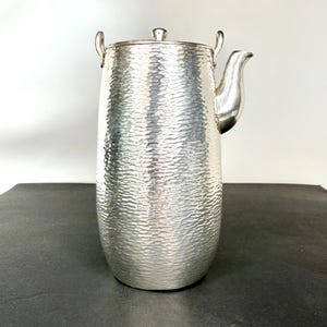 May include: A silver-colored, hammered-texture pitcher with a lid and a spout. The pitcher has two small handles on the top and a curved spout on the side. The surface has a dimpled texture, and the pitcher is sitting on a dark surface.