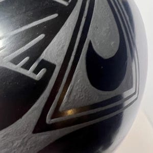 May include: A black ceramic vase with a glossy finish. The vase features a geometric design in a lighter gray color. The design includes angular shapes and curved lines, creating a striking contrast against the dark background.