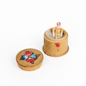 May include: Two wooden containers. One is a small, round box with a lid decorated with a painted flower design in red, blue, and yellow. The other is a cylindrical container with a small doll inside.