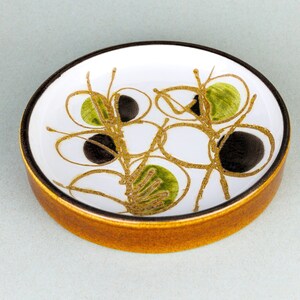 May include: A small, round ceramic dish with a brown rim and a white interior. The dish is decorated with a stylized design of black and green circles and lines.