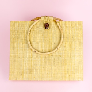 Puede incluir: Una bolsa de mano tejida de color marr&oacute;n claro con un asa circular de bamb&uacute;. La bolsa est&aacute; sobre un fondo rosa.