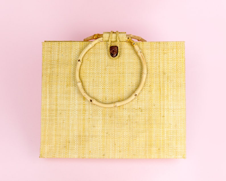 Puede incluir: Una bolsa de mano tejida de color marr&oacute;n claro con un asa circular de bamb&uacute;. La bolsa est&aacute; sobre un fondo rosa.