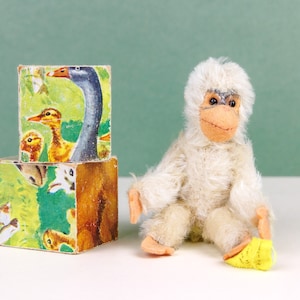 Peut inclure: Un singe en peluche blanc assis sur une surface blanche, tenant un jouet jaune. Le singe a un visage brun et des yeux noirs. Derrière le singe, il y a deux blocs de bois empilés avec des illustrations d'animaux colorés.