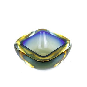 May include: A square-shaped glass ashtray with a gradient of colors. The rim is a mix of gold and yellow, transitioning to blue in the center. The base is a dark green color.