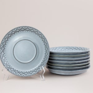 May include: A stack of eight blue ceramic saucers with a decorative pattern. The saucers are stacked on a light pink surface.