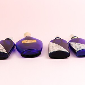 May include: Four cobalt blue glass bottles with white labels and silver lettering. The bottles are all different sizes and shapes. The labels on the bottles read "Eau de Cologne" and "Soie d'Orient".