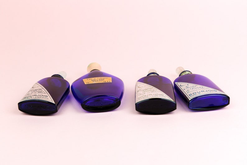 May include: Four cobalt blue glass bottles with white labels and silver lettering. The bottles are all different sizes and shapes. The labels on the bottles read "Eau de Cologne" and "Soie d'Orient".