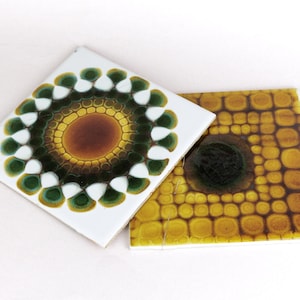 May include: Two square ceramic tiles with colorful designs. One tile features a white background with a green, yellow, and brown floral pattern. The other tile has a yellow background with a dark brown circular design.