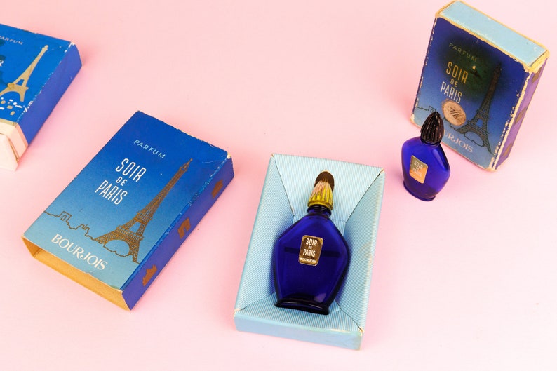Puede incluir: Tres cajas azules con letras doradas y un dise&ntilde;o de la Torre Eiffel dorada. Las cajas contienen botellas de perfume en miniatura etiquetadas como "Soir de Paris" de Bourjois.