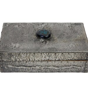 May include: A rectangular metal box with a textured, weathered appearance. The box is gray with a dark, iridescent stone on the lid. The box is riveted along the edges and has a hinged lid.