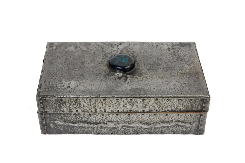 May include: A rectangular metal box with a textured, weathered appearance. The box is gray with a dark, iridescent stone on the lid. The box is riveted along the edges and has a hinged lid.