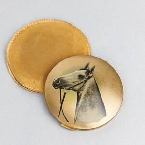 May include: A vintage gold-toned compact with a horse portrait on the lid, next to a matching round, gold-coloured pouch. The horse is depicted in a grayscale drawing. The compact is open, revealing the mirror and powder area.