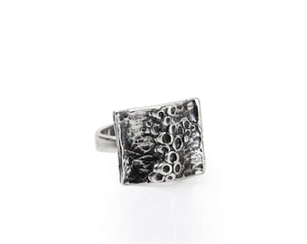 May include: A silver ring with a square-shaped face featuring a textured design. The ring is made of metal and has a simple band.