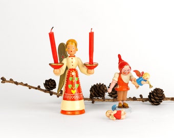 May include: A wooden angel figurine with red candles, a wooden gnome figurine holding a wooden angel figurine, and a wooden angel figurine lying on the ground. All figurines are painted in bright colors and have a whimsical, folk art style.