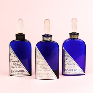 May include: Three blue glass bottles with silver labels and clear glass stoppers. The labels have the text "Evening in Paris", "Soir de Paris", and "Bourjois Paris London" on them.