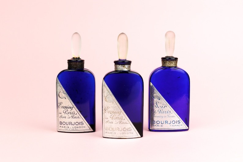 May include: Three blue glass bottles with silver labels and clear glass stoppers. The labels have the text "Evening in Paris", "Soir de Paris", and "Bourjois Paris London" on them.