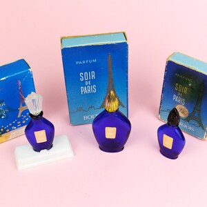 Puede incluir: Tres frascos en miniatura de perfume Soir de Paris, cada uno con un frasco de vidrio azul y una tapa dorada. Los frascos se muestran sobre una superficie rosa con sus cajas originales.