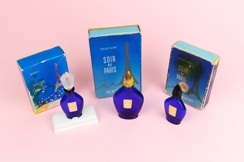 Puede incluir: Tres frascos en miniatura de perfume Soir de Paris, cada uno con un frasco de vidrio azul y una tapa dorada. Los frascos se muestran sobre una superficie rosa con sus cajas originales.