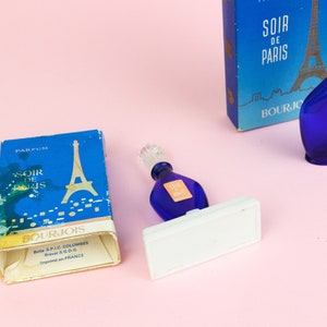 Puede incluir: Un frasco de perfume azul de Bourjois Soir de Paris con un tap&oacute;n dorado. El frasco est&aacute; sobre un soporte blanco. El frasco est&aacute; al lado de una caja del mismo perfume con una imagen de la Torre Eiffel.