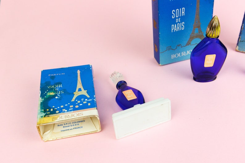 Puede incluir: Un frasco de perfume azul de Bourjois Soir de Paris con un tap&oacute;n dorado. El frasco est&aacute; sobre un soporte blanco. El frasco est&aacute; al lado de una caja del mismo perfume con una imagen de la Torre Eiffel.