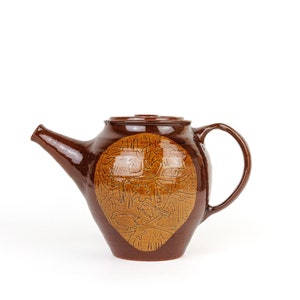 May include: A brown ceramic teapot with a carved design on the side. The design is a light brown colour and features a stylised tree and birds.