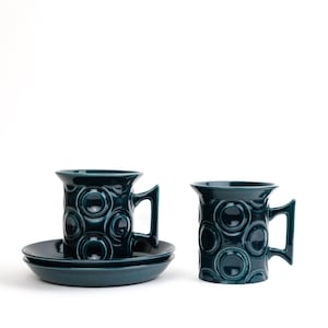 May include: Two teal blue ceramic mugs with a circular pattern and matching saucers. The mugs have a rounded handle.