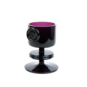 May include: A black glass goblet with a purple interior and a decorative seal on the side. The goblet sits on a three-tiered base.