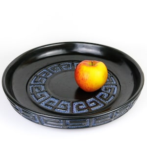 Peut inclure: Un plat de service en céramique noire avec un motif géométrique bleu et blanc autour du bord. Une pomme se trouve au centre du plat.