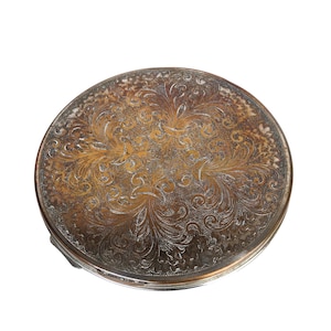 May include: A round, antique silver compact with an ornate, engraved floral design. The compact has a tarnished, aged appearance, with a mix of silver and gold tones. The detailed pattern covers the entire top surface.