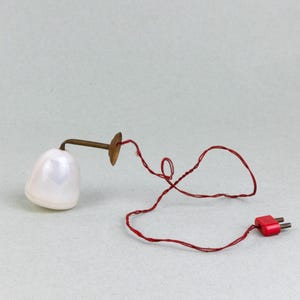 May include: A white ceramic lamp with a red cord and a brass base. The lamp has a simple design and is perfect for a bedside table or desk.