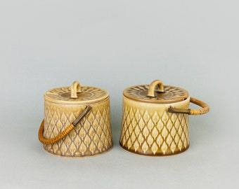 May include: Two ceramic lidded jars with brown and white leaf patterns and wooden handles. The jars are on a light gray background.