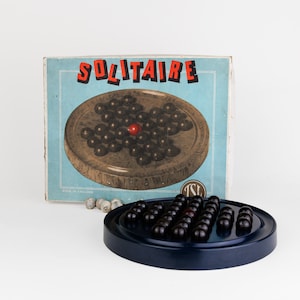 May include: A vintage solitaire game with a blue board and black marbles. The game is in its original box, which has a blue background and the word "SOLITAIRE" printed in red letters.
