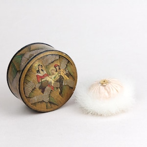 May include: A vintage round powder box with a decorative lid depicting a religious scene in gold, red, and green. A fluffy, pale pink powder puff with white trim is next to the box. The box has a black rim.